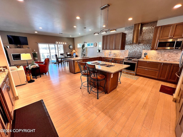 kitchen featuring a kitchen breakfast bar, decorative light fixtures, light wood-type flooring, wall chimney range hood, and stainless steel appliances