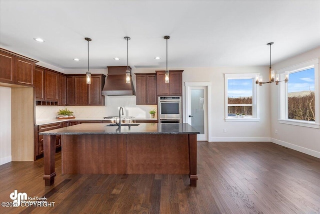 kitchen with double oven, decorative light fixtures, custom range hood, and an island with sink