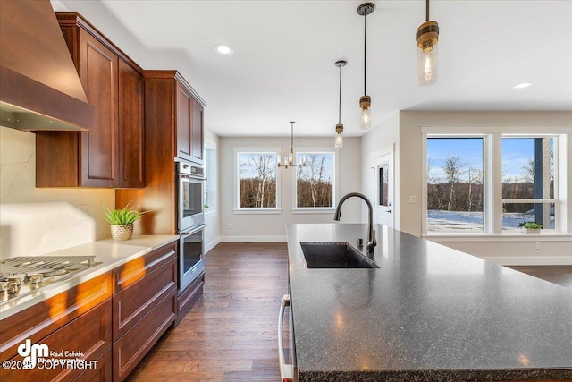 kitchen with sink, a center island with sink, appliances with stainless steel finishes, pendant lighting, and wall chimney range hood