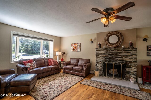 living room featuring a textured ceiling, wood-type flooring, a stone fireplace, baseboard heating, and ceiling fan