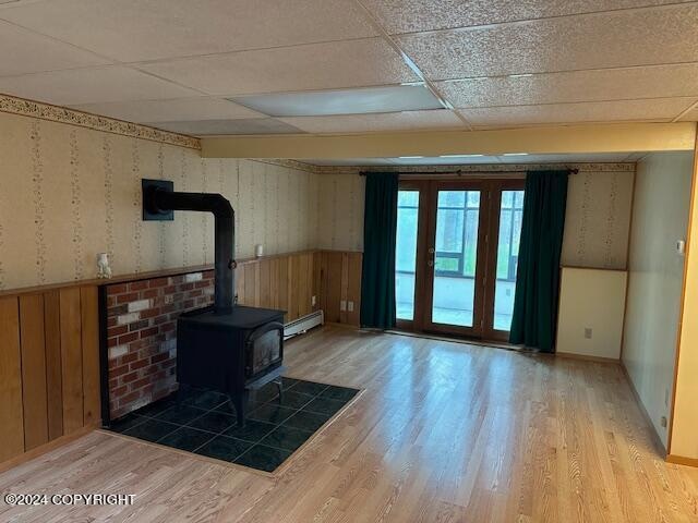 living room featuring light wood-type flooring, a wood stove, a paneled ceiling, wood walls, and baseboard heating