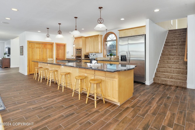 kitchen with stainless steel appliances, a center island, decorative light fixtures, dark stone counters, and light brown cabinets