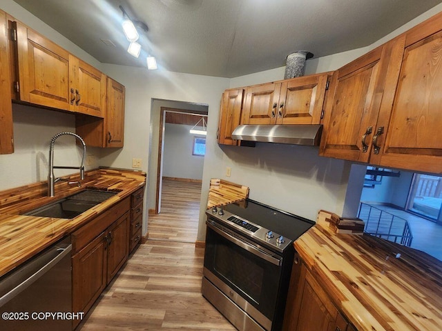 kitchen featuring stainless steel appliances, brown cabinetry, a sink, butcher block countertops, and under cabinet range hood