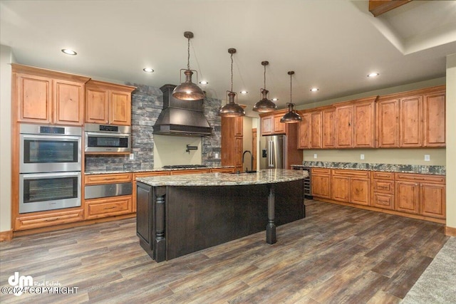 kitchen with dark hardwood / wood-style flooring, appliances with stainless steel finishes, a kitchen island with sink, light stone counters, and custom range hood