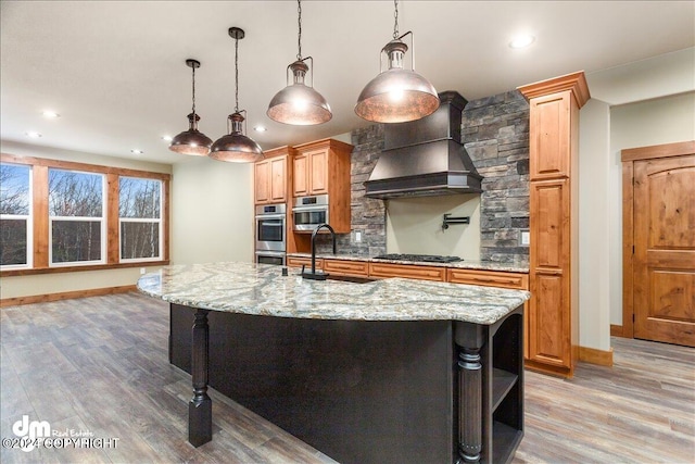 kitchen featuring stainless steel double oven, a kitchen island with sink, light stone countertops, custom range hood, and hardwood / wood-style floors