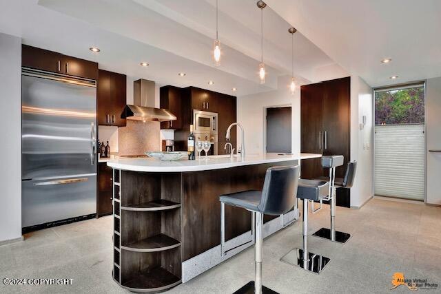 kitchen featuring built in appliances, dark brown cabinetry, light countertops, wall chimney range hood, and open shelves