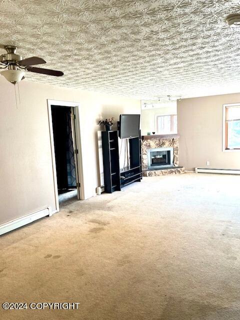 unfurnished living room featuring a baseboard radiator, ceiling fan, a textured ceiling, carpet flooring, and a stone fireplace