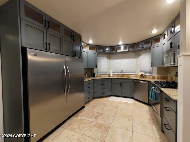 kitchen featuring light tile patterned floors, glass insert cabinets, light stone counters, stainless steel appliances, and a sink