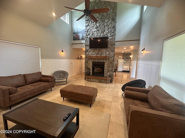 tiled living area featuring a wainscoted wall, a stone fireplace, a towering ceiling, and a ceiling fan