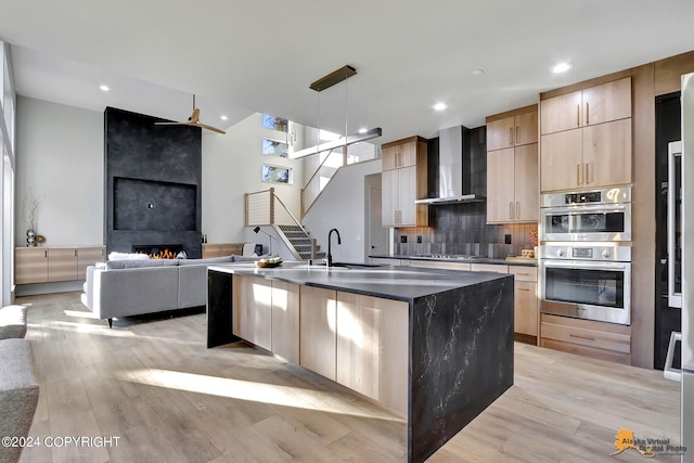 kitchen with wall chimney exhaust hood, light brown cabinets, a large fireplace, light hardwood / wood-style flooring, and decorative light fixtures