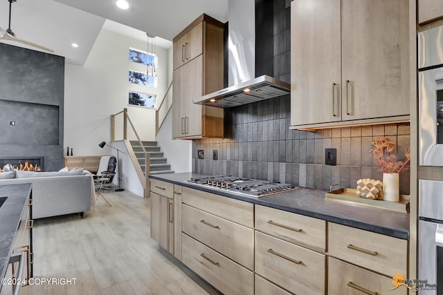 kitchen featuring tasteful backsplash, wall chimney exhaust hood, stainless steel gas stovetop, a large fireplace, and light wood-type flooring