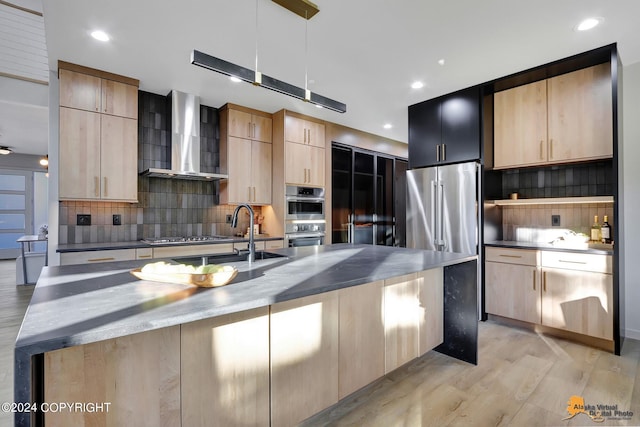 kitchen with tasteful backsplash, a center island with sink, wall chimney range hood, appliances with stainless steel finishes, and light hardwood / wood-style floors