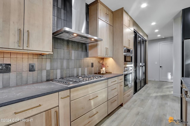 kitchen with light wood-type flooring, wall chimney range hood, backsplash, dark stone countertops, and light brown cabinetry