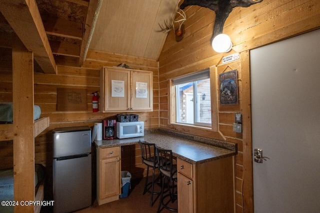 kitchen featuring light brown cabinetry, stainless steel fridge, vaulted ceiling, and wood walls