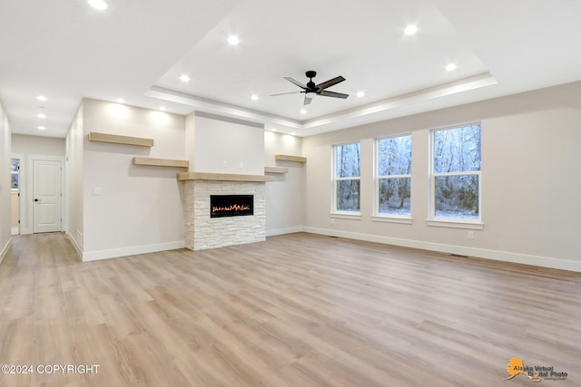 unfurnished living room with ceiling fan, a stone fireplace, a tray ceiling, and light hardwood / wood-style floors