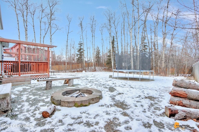 yard covered in snow with a trampoline, a wooden deck, and an outdoor fire pit