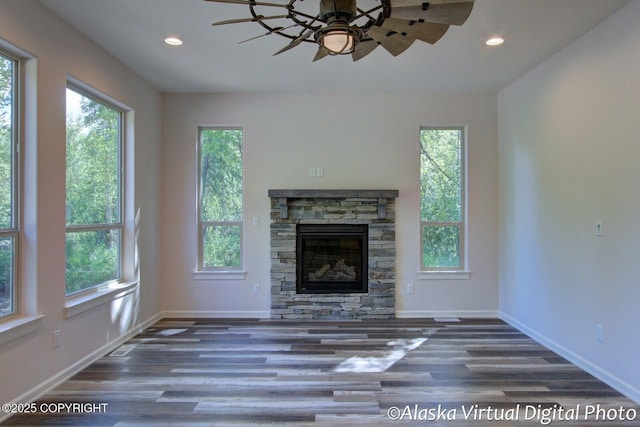 unfurnished living room with ceiling fan, a stone fireplace, and dark wood-type flooring