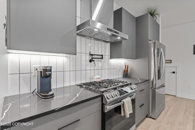 kitchen featuring appliances with stainless steel finishes, light wood-type flooring, tasteful backsplash, wall chimney exhaust hood, and dark stone countertops