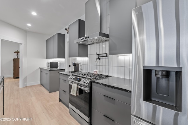 kitchen with wall chimney range hood, gray cabinets, appliances with stainless steel finishes, tasteful backsplash, and light hardwood / wood-style floors