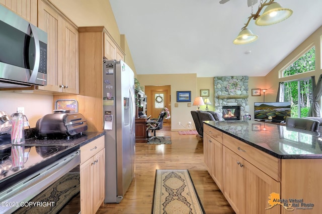 kitchen with vaulted ceiling, light brown cabinetry, light hardwood / wood-style floors, appliances with stainless steel finishes, and a stone fireplace