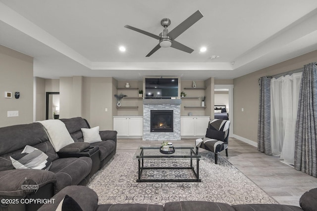 living room featuring light hardwood / wood-style floors, built in shelves, a tray ceiling, and a stone fireplace