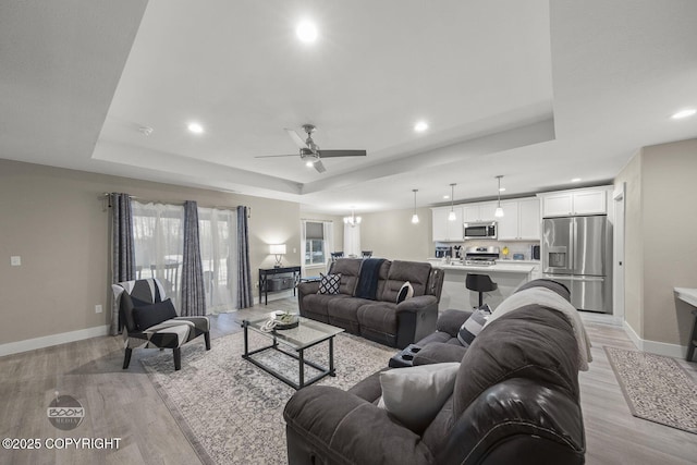 living room with ceiling fan with notable chandelier, a tray ceiling, and light wood-type flooring