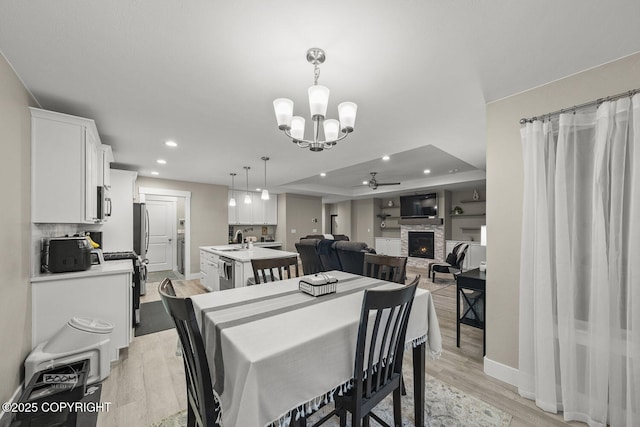 dining area with light hardwood / wood-style flooring, a raised ceiling, sink, a fireplace, and ceiling fan with notable chandelier