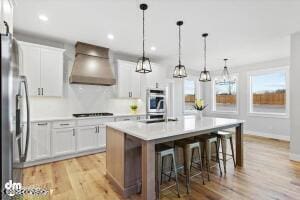 kitchen with white cabinetry, a kitchen island with sink, wall chimney range hood, decorative light fixtures, and stainless steel appliances