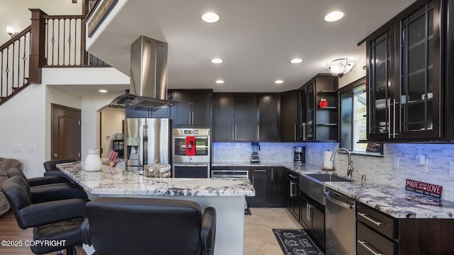 kitchen with a kitchen bar, sink, island exhaust hood, stainless steel appliances, and light stone countertops