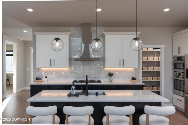 kitchen featuring white cabinetry, tasteful backsplash, hanging light fixtures, an island with sink, and wall chimney range hood