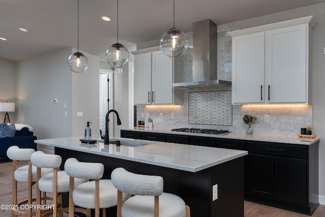 kitchen featuring wall chimney range hood, sink, a kitchen island with sink, white cabinetry, and stainless steel gas cooktop