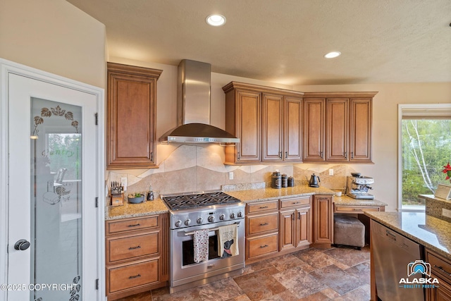 kitchen with wall chimney range hood, appliances with stainless steel finishes, decorative backsplash, light stone counters, and brown cabinets