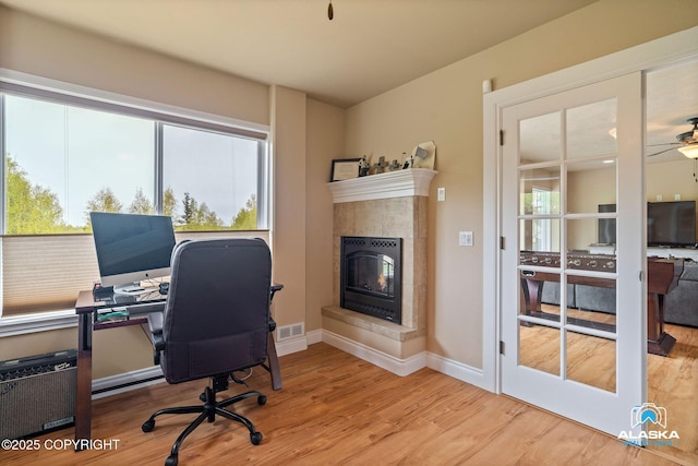 office with light wood-style flooring, a tiled fireplace, a ceiling fan, and baseboards