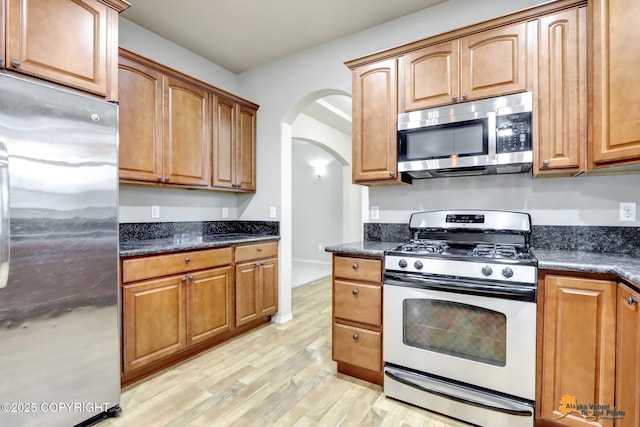 kitchen with arched walkways, stainless steel appliances, brown cabinets, and light wood-style floors