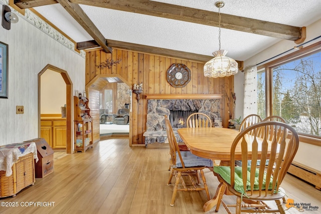 dining area featuring light hardwood / wood-style flooring, a stone fireplace, lofted ceiling with beams, a textured ceiling, and wooden walls