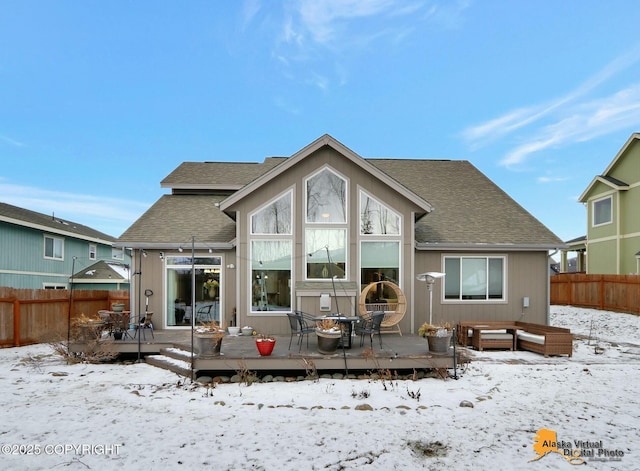 snow covered house with a shingled roof, fence, and a wooden deck