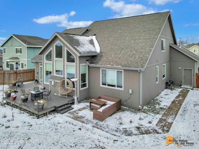 snow covered property with a deck, roof with shingles, and fence