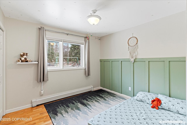 bedroom featuring a decorative wall, a baseboard heating unit, wood finished floors, baseboards, and wainscoting