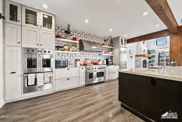 kitchen featuring high end appliances, open shelves, a sink, wall chimney exhaust hood, and light wood-type flooring