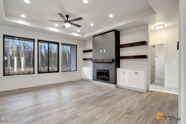 unfurnished living room with a lit fireplace, a tray ceiling, light wood-style flooring, and baseboards