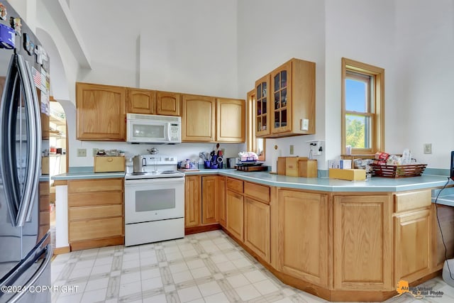 kitchen featuring light countertops, a towering ceiling, glass insert cabinets, white appliances, and a peninsula