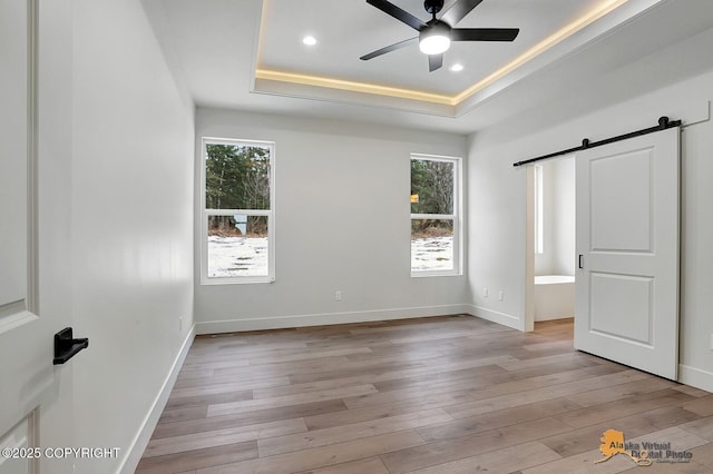 unfurnished bedroom featuring a tray ceiling, a barn door, multiple windows, and light wood-style flooring