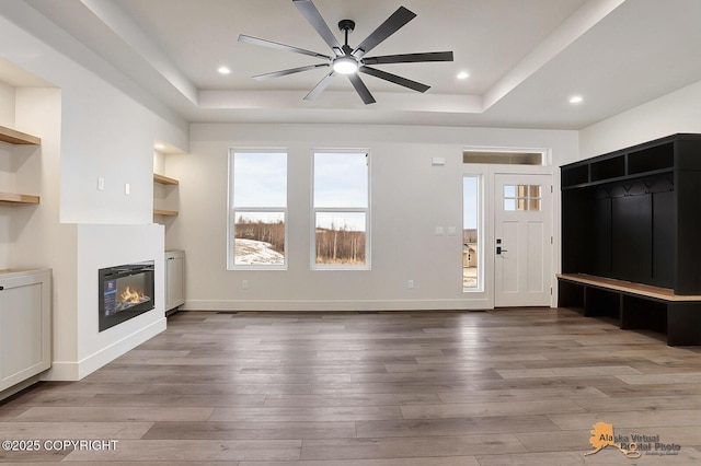 unfurnished living room featuring a glass covered fireplace, a raised ceiling, baseboards, and wood finished floors