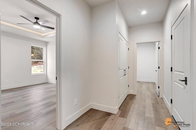 corridor featuring light wood-style flooring, baseboards, a tray ceiling, and recessed lighting