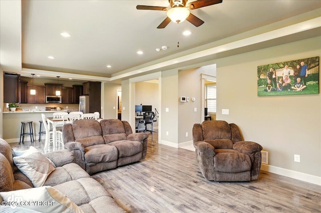 living area with ceiling fan, light wood-type flooring, a tray ceiling, recessed lighting, and baseboards