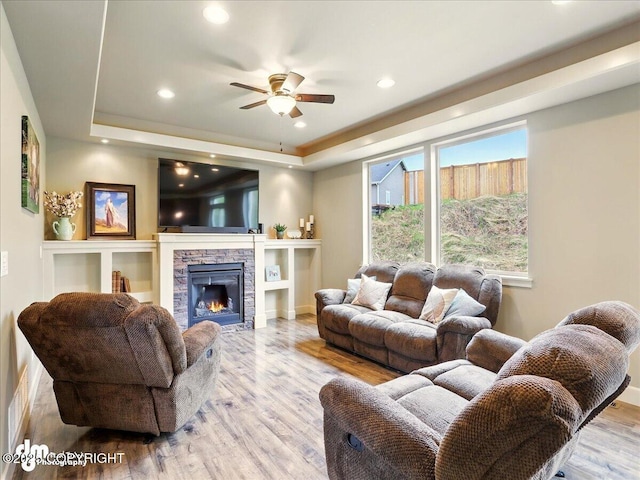living area featuring a tray ceiling, wood finished floors, ceiling fan, a stone fireplace, and recessed lighting