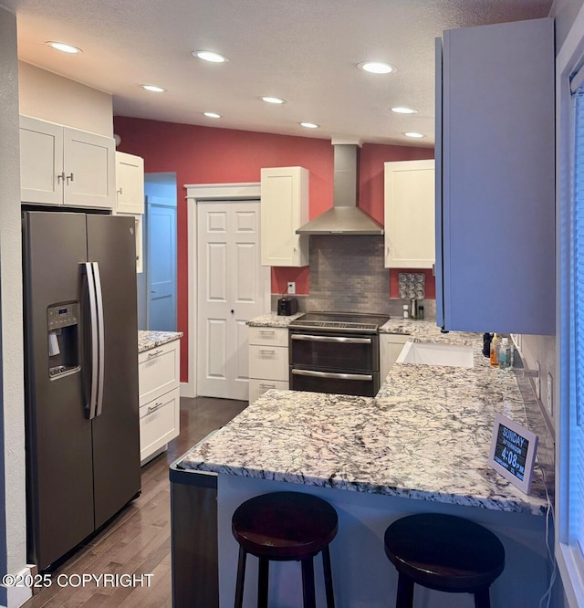 kitchen featuring wall chimney exhaust hood, appliances with stainless steel finishes, a breakfast bar area, a peninsula, and light stone countertops