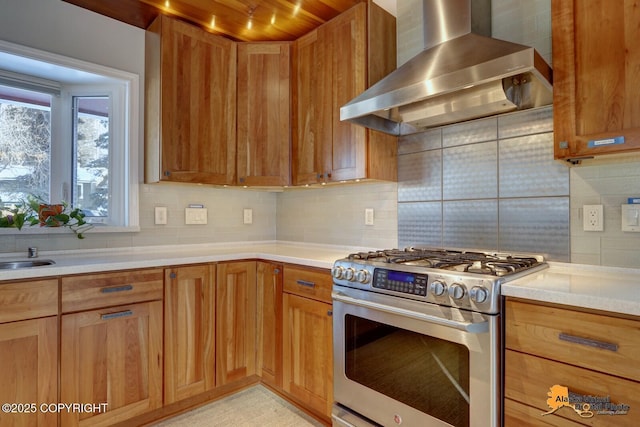 kitchen with stainless steel gas stove, backsplash, light countertops, wall chimney range hood, and brown cabinetry