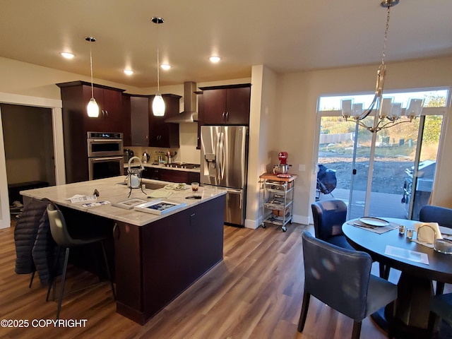 kitchen with stainless steel appliances, wood finished floors, hanging light fixtures, wall chimney range hood, and dark brown cabinets