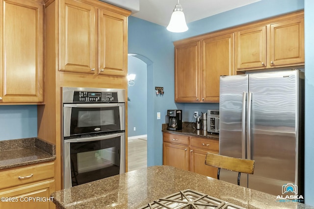 kitchen featuring dark stone countertops, baseboards, arched walkways, light brown cabinetry, and appliances with stainless steel finishes